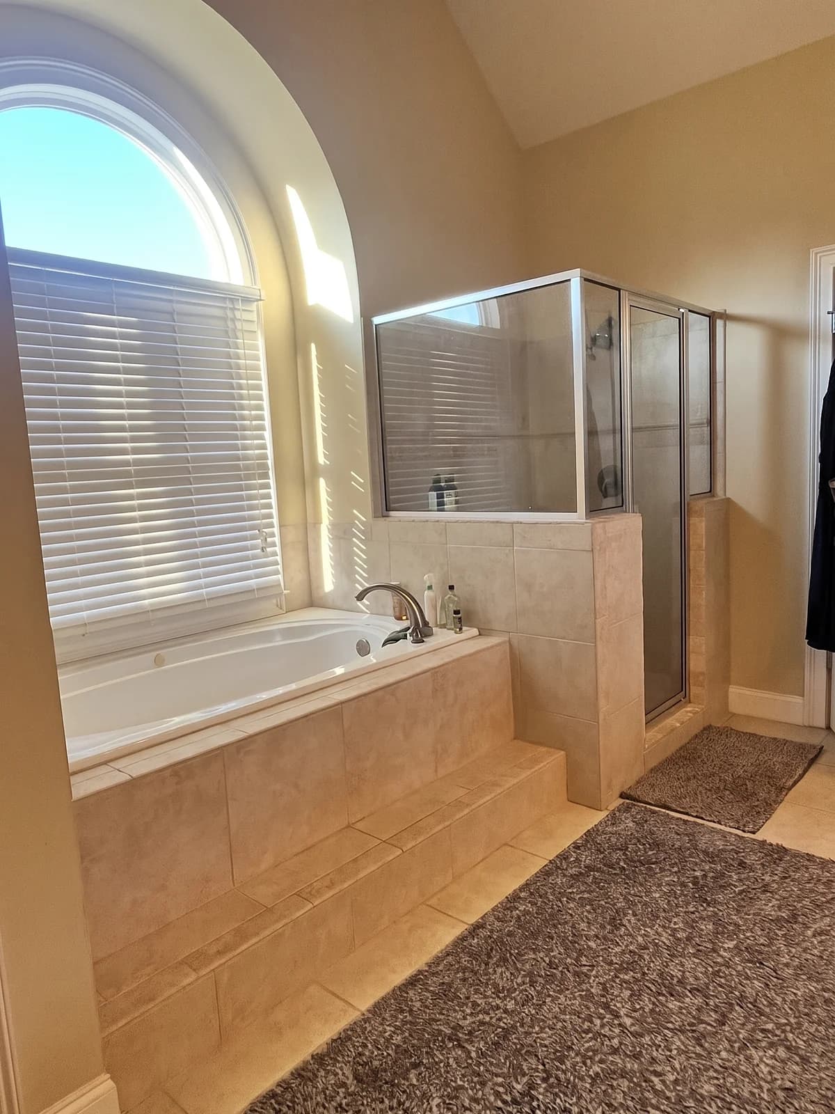 Before: drop-in tub in a beige tile deck with steps, under an arched window, next to a framed glass shower