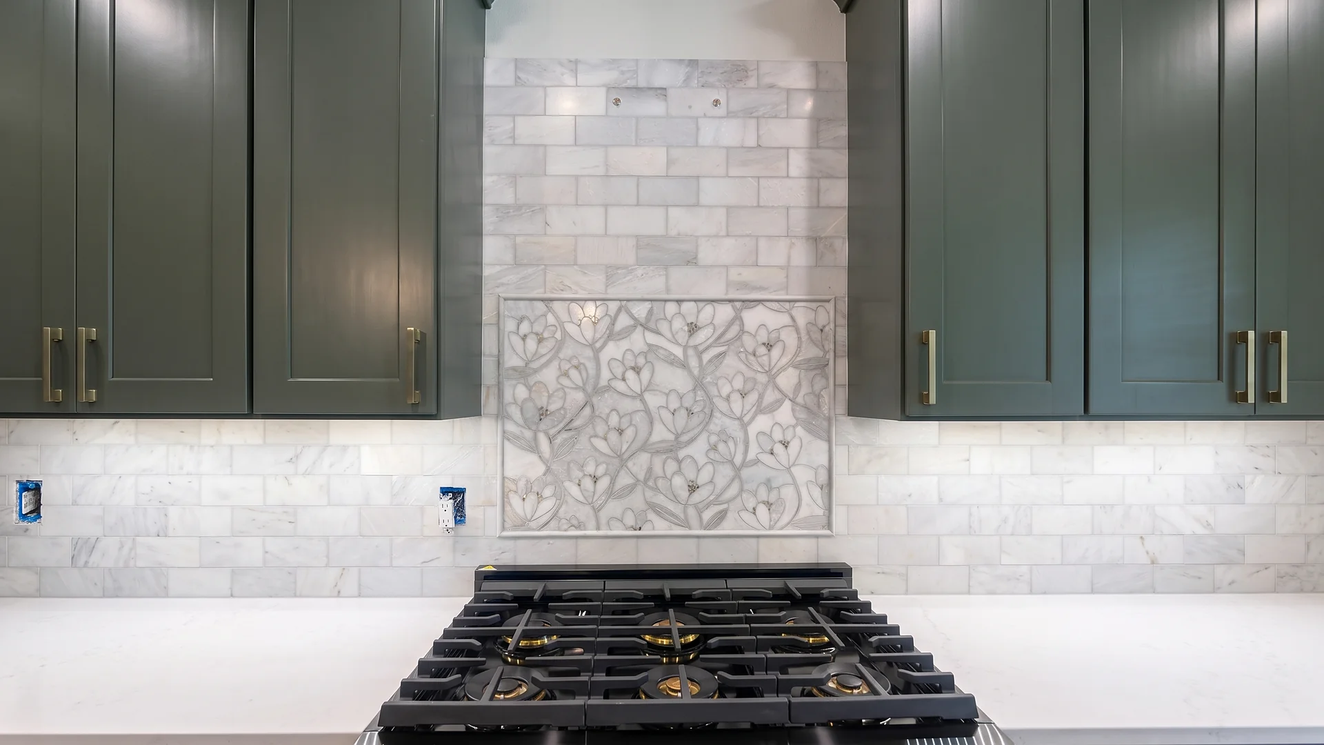 Marble-look subway backsplash with a floral mosaic inset above a gas range, between green shaker cabinets