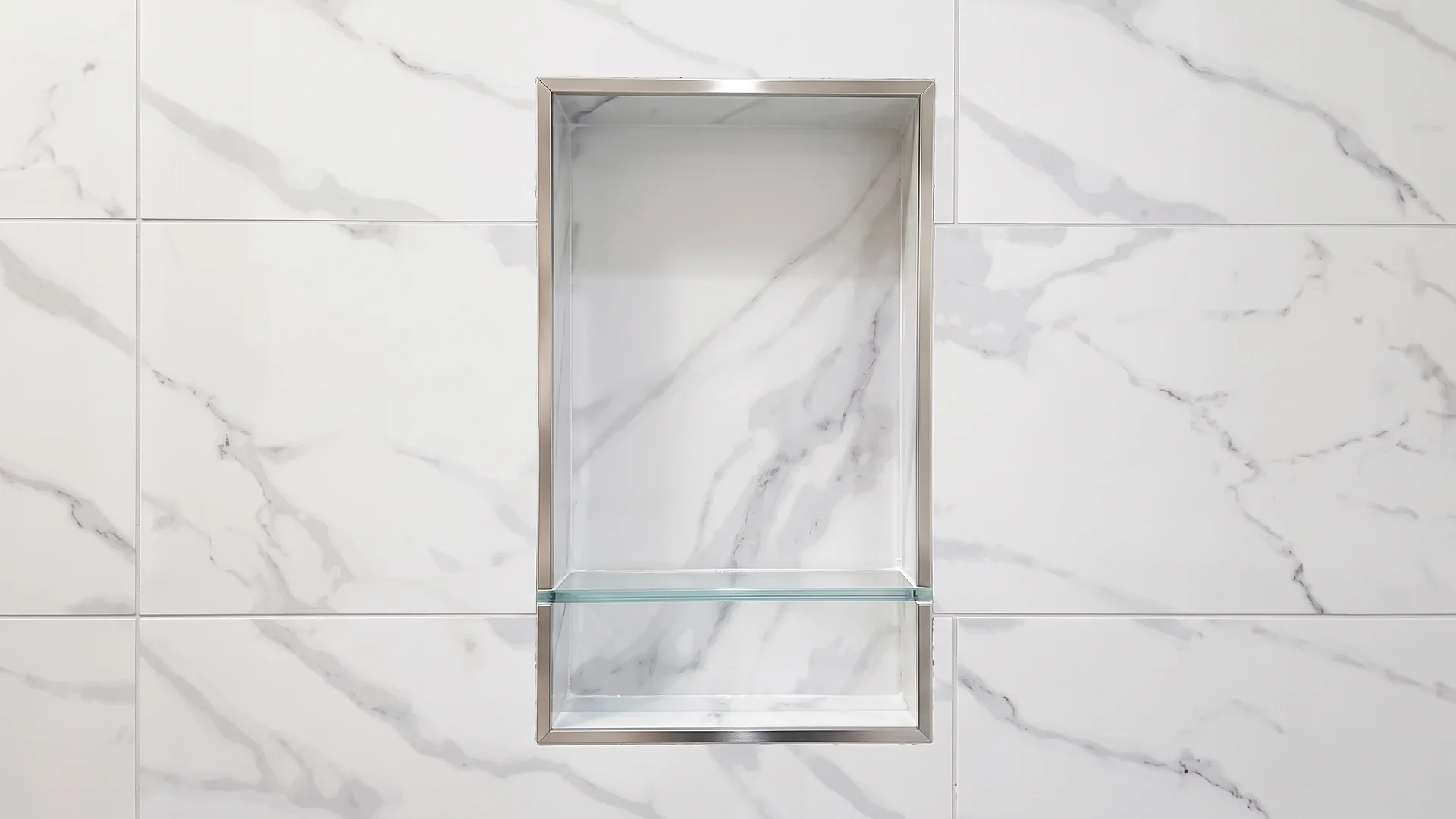 Recessed shower niche with a glass shelf set in marble-look wall tile with tight grout lines